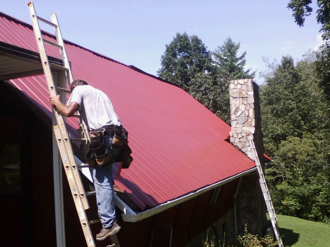 Roofer on a ladder beside a red metal roof and stone chimney — Soffit & Fascia Repair by Roofing Brothers in Monmouth